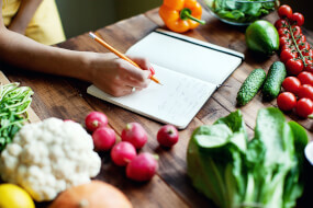 Woman writing in a notebook a meal plan with fresh vegetables on the table