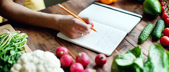 Woman writing in a notebook a meal plan with fresh vegetables on the table