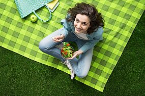 Woman on a picnic blanket eating a salad looking up at the camera